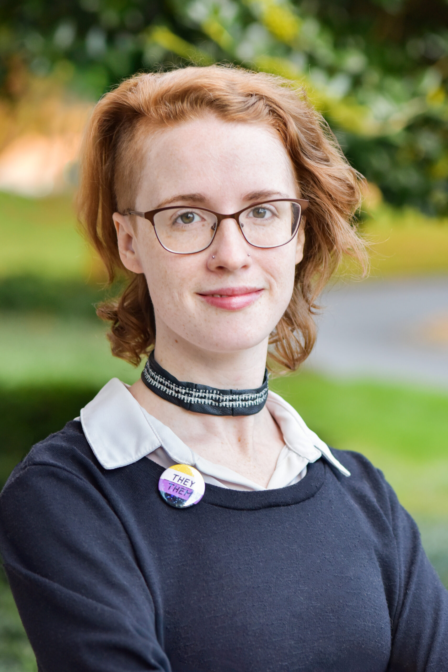 A white person in their 30s with red hair in a side shave style wearing glasses stands outdoors, wearing a black sweater over a white collared shirt, a black fabric choker necklace, and a pin that reads “they/them.” The background shows greenery and a blurred pathway.